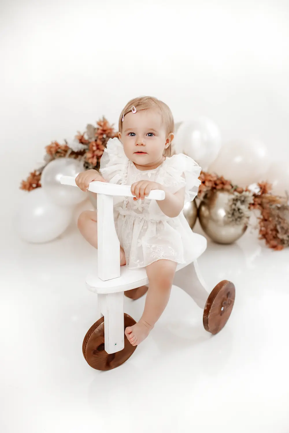 Uśmiechnięty maluch w białej sukience siedzi na drewnianym trójkołowym rowerku, otoczony białymi balonami i suszonymi kwiatami. Scena jest jasna i radosna.