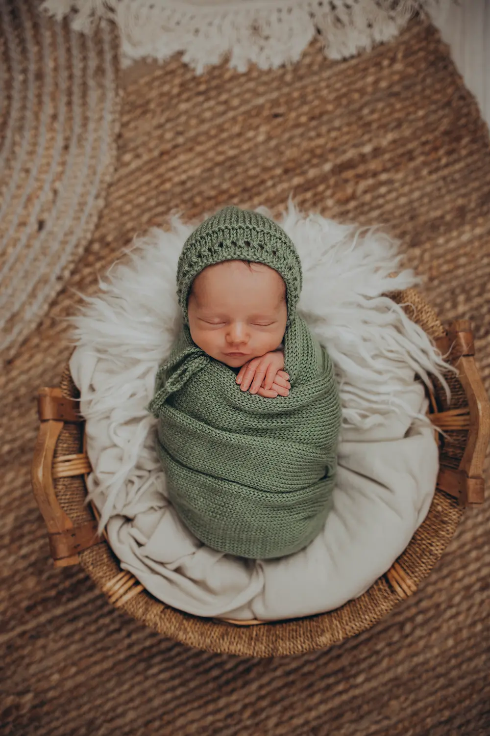Noworodek owinięty w zielony dziany kocyk i czapeczkę, śpiący w wiklinowym koszu wyłożonym białymi kocami na fakturowanym dywanie.