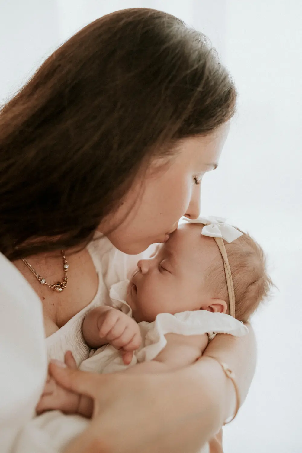 Matka delikatnie przytula swoje niemowlę, otulona miękkimi tkaninami w ciepłej, pełnej czułości chwili w jasnym, miękko oświetlonym pokoju.