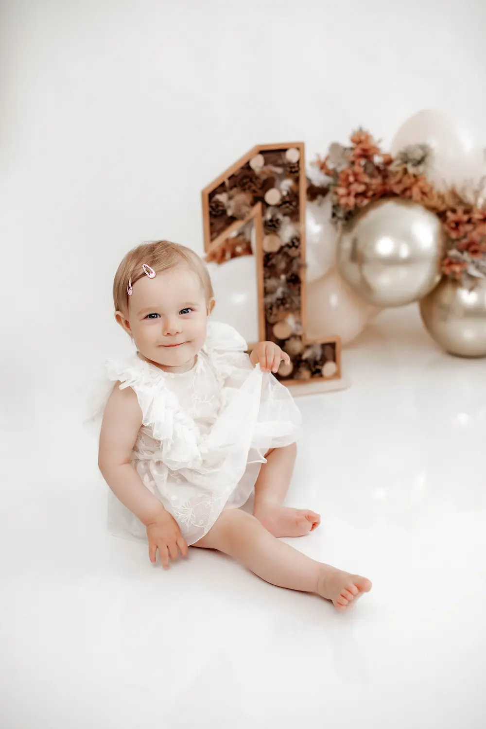Maluch w białej sukience z falbankami siedzi na błyszczącej powierzchni, uśmiechając się obok dekoracyjnej drewnianej cyfry jeden i pastelowych balonów.
