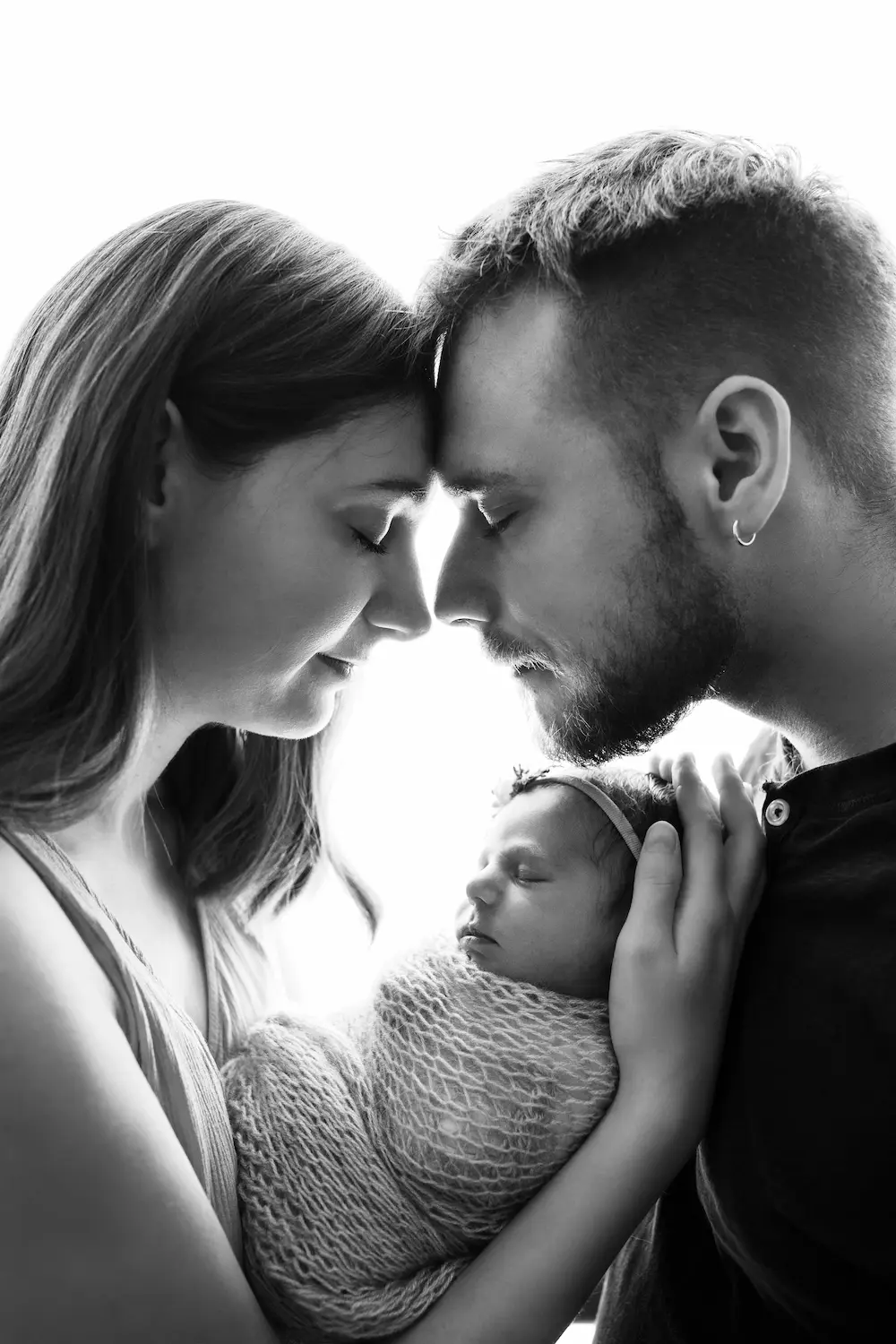 Para delikatnie się przytula, wspólnie trzymając owiniętego noworodka, uchwycona w miękkim czarno-białym portrecie.