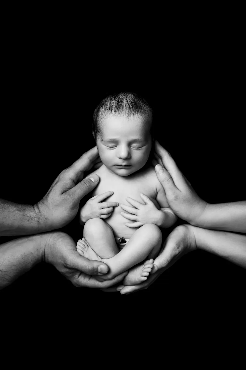 Noworodek siedzi otoczony wieloma dłońmi na tle głębokiej czerni, symbolizując miłość i troskę.