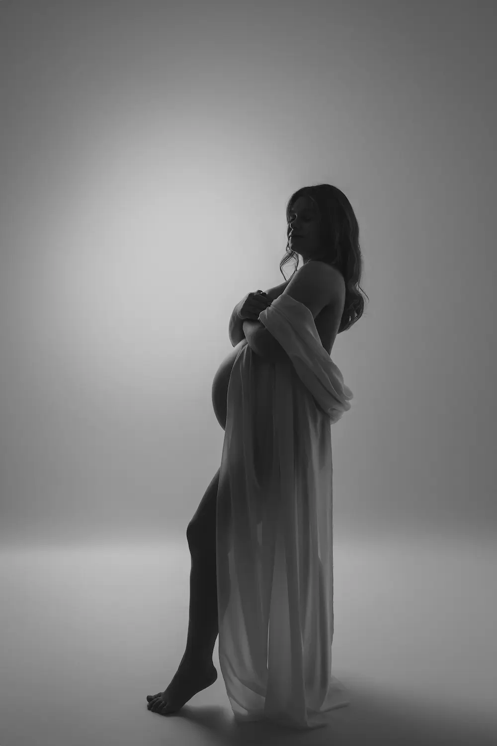 Sylwetka kobiety w ciąży owiniętej lejącą się tkaniną, stojącej boso. Oświetlenie tworzy miękką, spokojną atmosferę podkreślającą jej kształt.